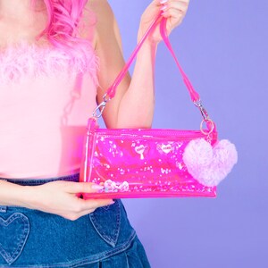 May include: A clear pink handbag with a matching strap and a fluffy pink heart-shaped charm. The bag contains small, decorative elements and the word "XOXO" is printed on the front. The bag is held against a purple background.