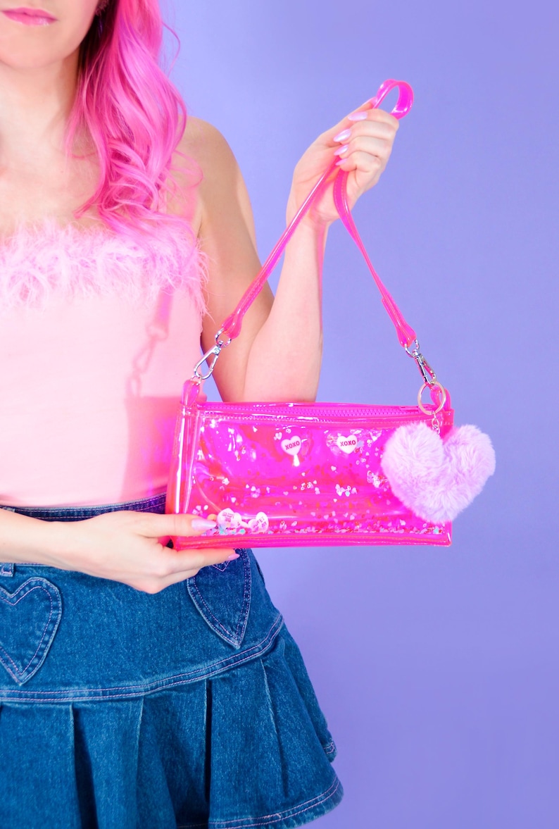 May include: A clear pink handbag with a matching strap and a fluffy pink heart-shaped charm. The bag contains small, decorative elements and the word "XOXO" is printed on the front. The bag is held against a purple background.
