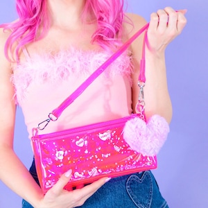 May include: A vibrant pink crossbody bag with a clear front, adorned with white heart and "XOXO" motifs. It has a matching strap and a fluffy pink heart-shaped charm. The bag is held up by a person.