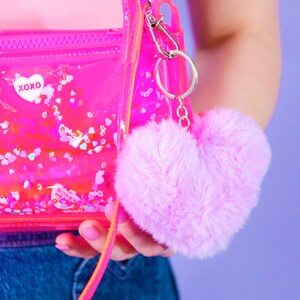 May include: A transparent pink purse with a zip and a pink strap. The purse has the text "XOXO" and a fluffy, heart-shaped pink keychain. The purse contains confetti.