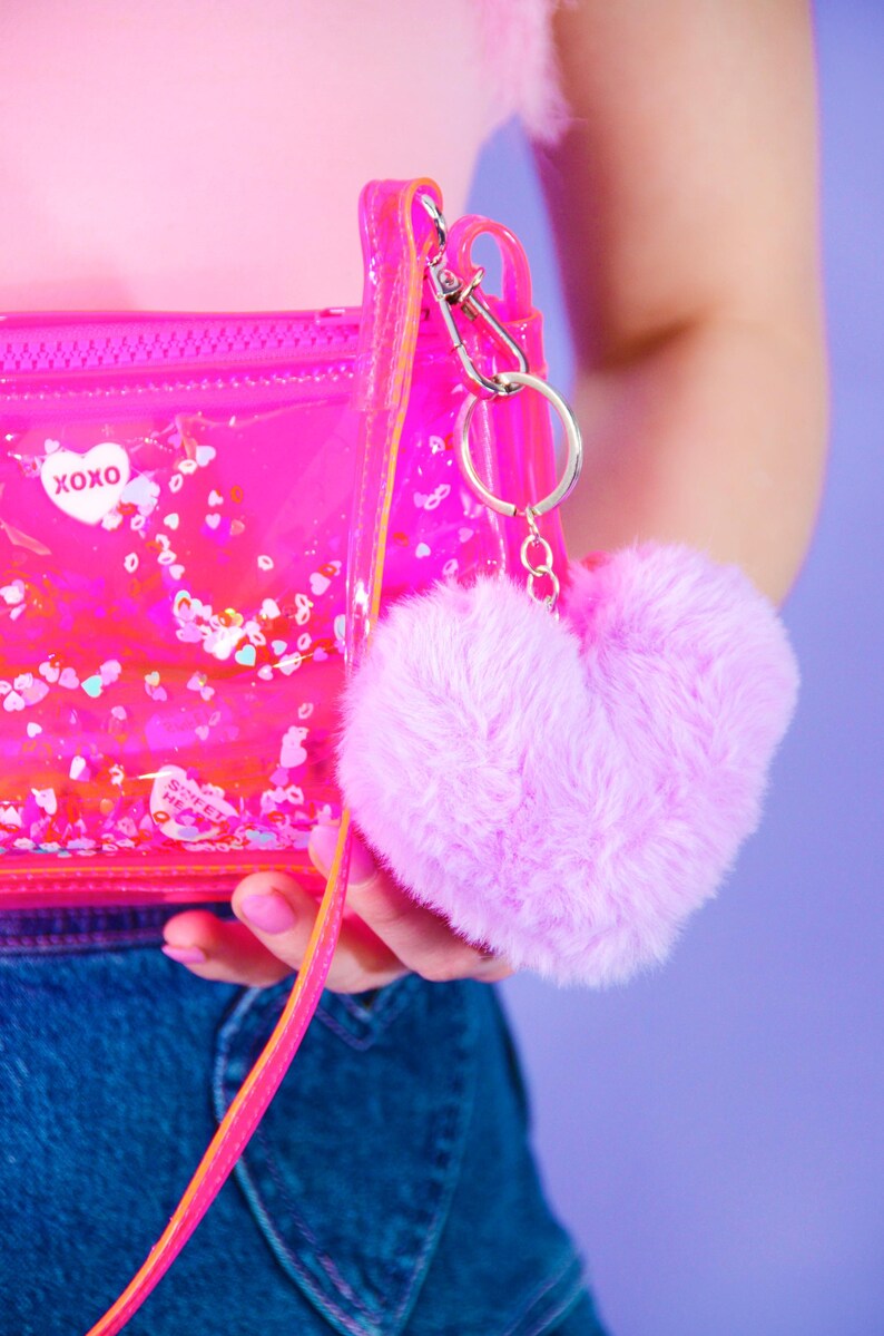 May include: A transparent pink purse with a zip and a pink strap. The purse has the text "XOXO" and a fluffy, heart-shaped pink keychain. The purse contains confetti.