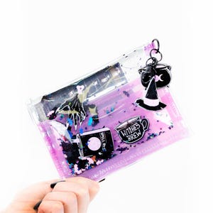 May include: A clear, rectangular pouch filled with purple liquid and glitter, featuring graphics of a hand, a spell book, and a mug labeled Witches Brew. A black metal keychain with a cauldron and witch hat charm is attached to the zipper.