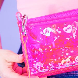 May include: A clear, pink zippered pouch with a silver clasp and a pink and yellow strap. The pouch is filled with heart-shaped confetti and candy hearts with the words "BE MINE" and "XOXO".