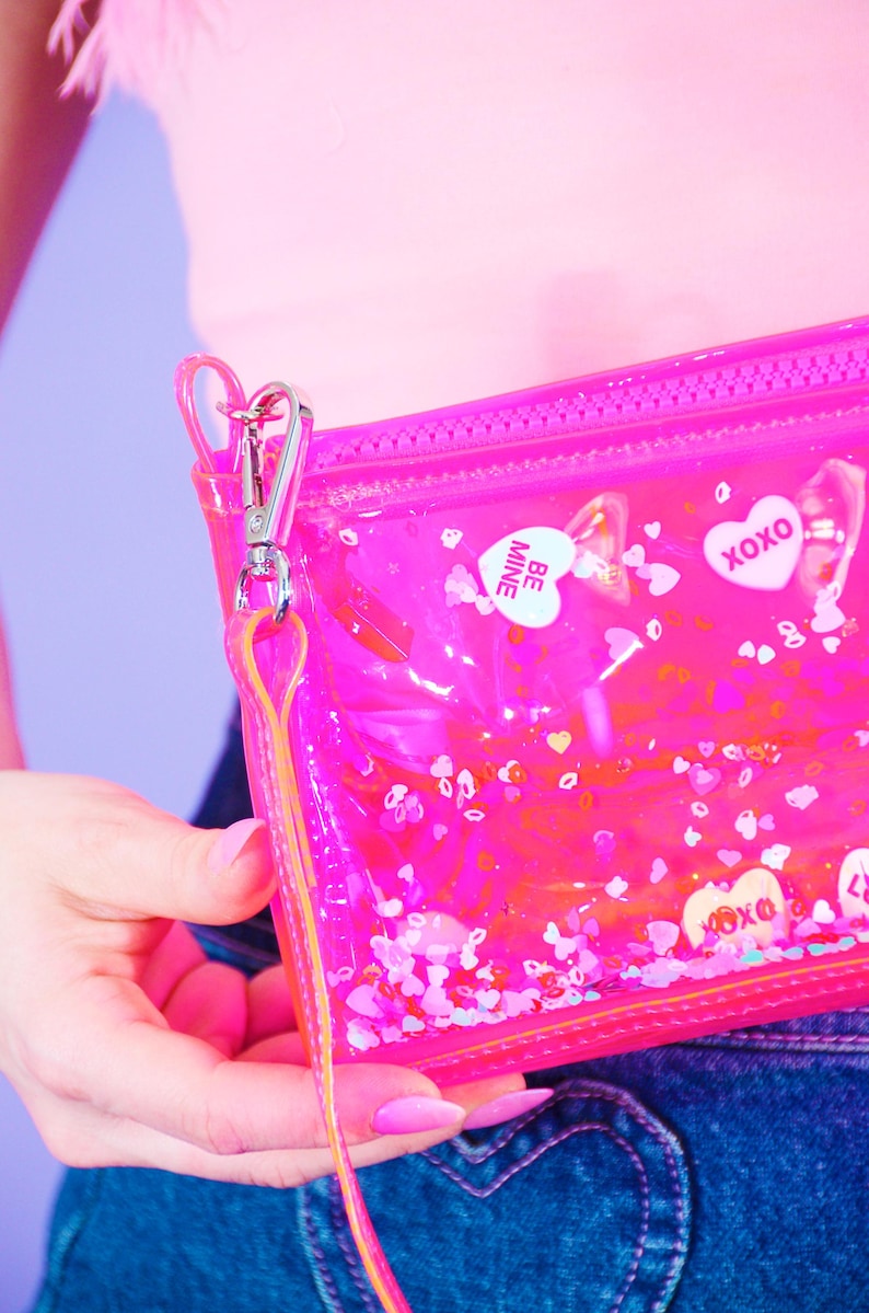 May include: A clear, pink zippered pouch with a silver clasp and a pink and yellow strap. The pouch is filled with heart-shaped confetti and candy hearts with the words "BE MINE" and "XOXO".