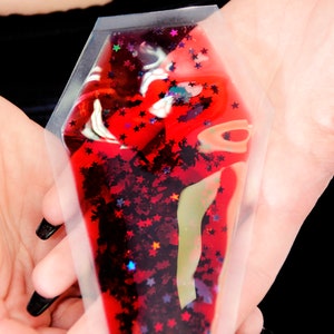 May include: A coffin-shaped, clear plastic pouch filled with red liquid and star-shaped glitter. The pouch is held in open hands against a black background. The glitter includes red, orange, and blue stars.