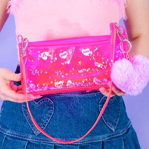 May include: A clear pink rectangular purse with a zip, a long strap, and a fluffy pink heart-shaped keyring. The purse contains floating confetti and heart-shaped stickers with the text "XOXO" and "Be Mine".