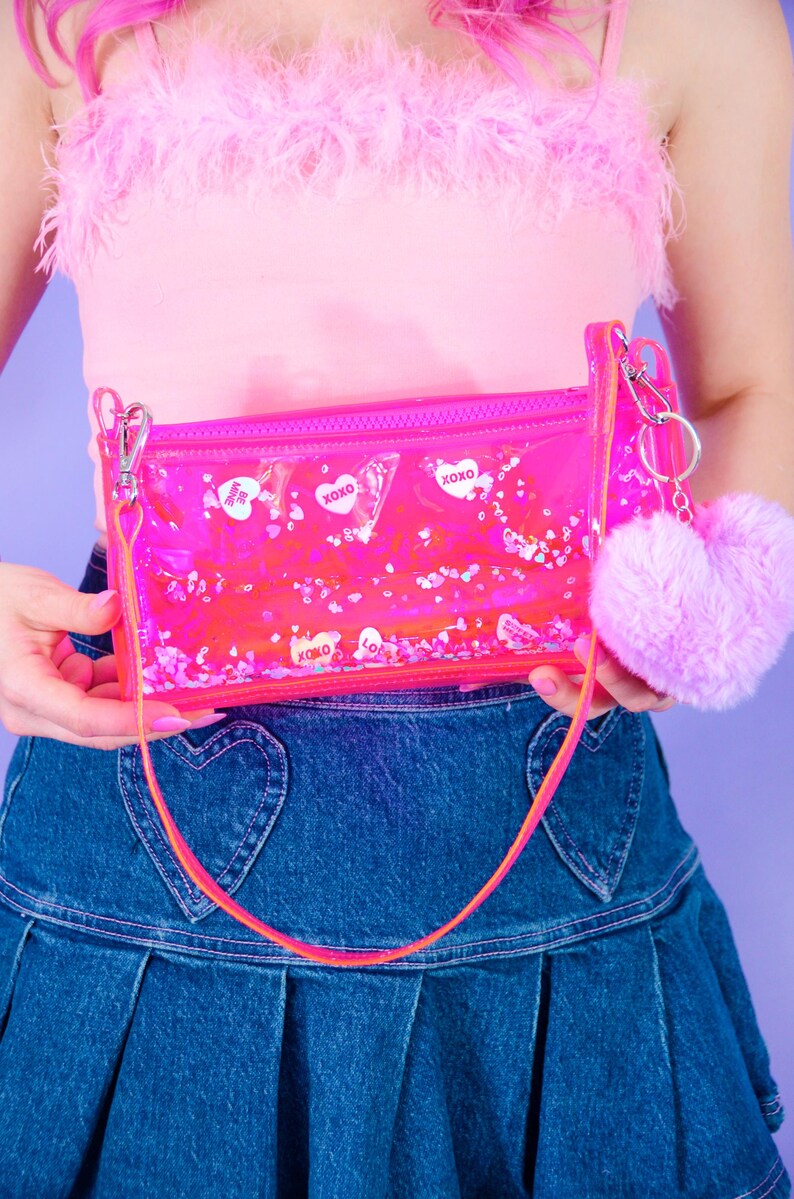 May include: A clear pink rectangular purse with a zip, a long strap, and a fluffy pink heart-shaped keyring. The purse contains floating confetti and heart-shaped stickers with the text "XOXO" and "Be Mine".