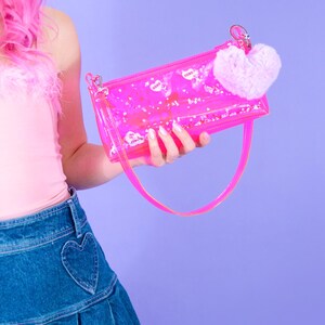 May include: A clear pink handbag with a matching strap and a fluffy pink heart charm. The bag contains small white confetti and heart-shaped stickers with the word "xoxo". The bag is held against a purple background.