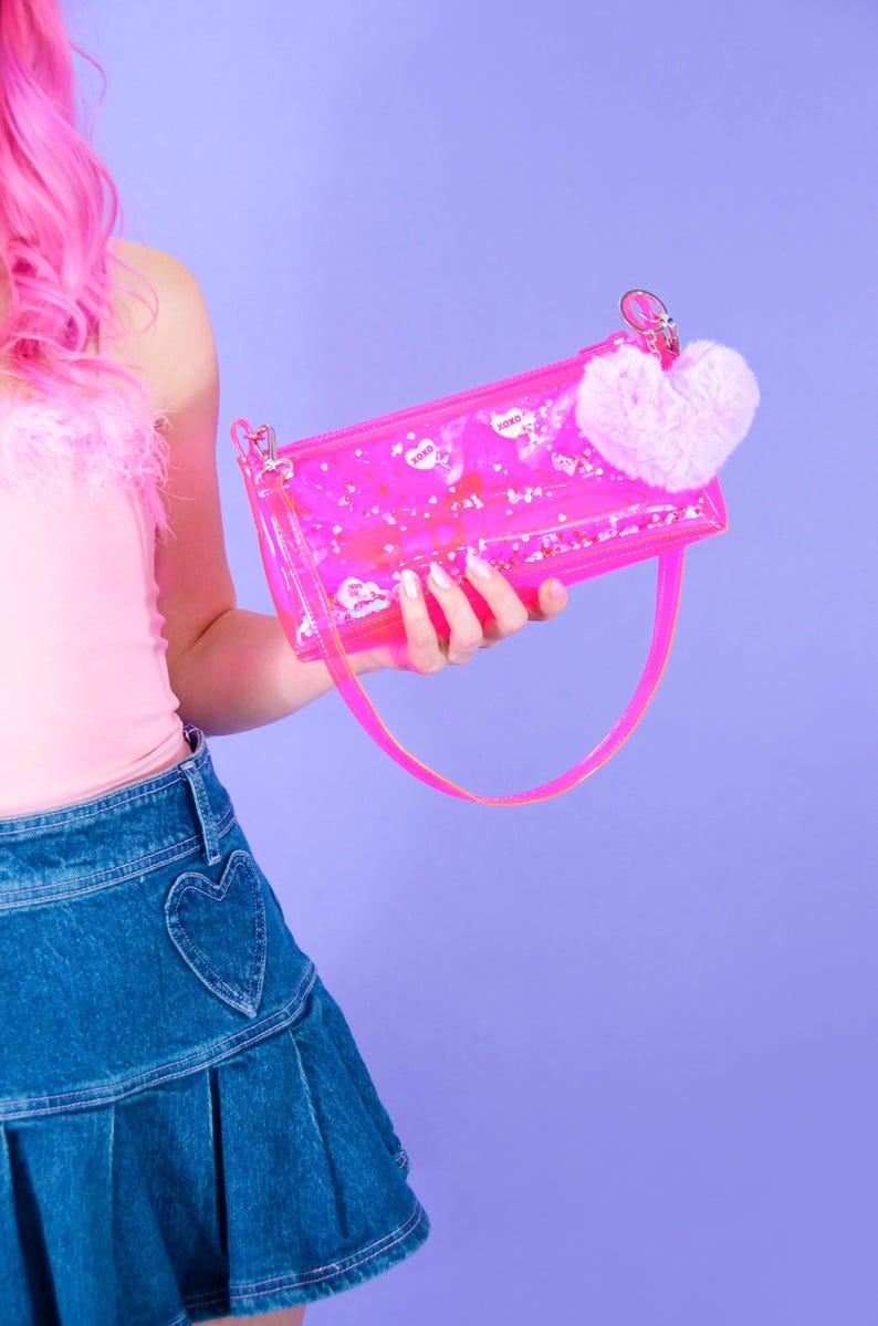 May include: A clear pink handbag with a matching strap and a fluffy pink heart charm. The bag contains small white confetti and heart-shaped stickers with the word "xoxo". The bag is held against a purple background.