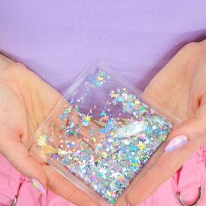May include: A clear plastic pouch filled with iridescent glitter in the shape of stars and crescent moons. The pouch is held in the hands of a person.