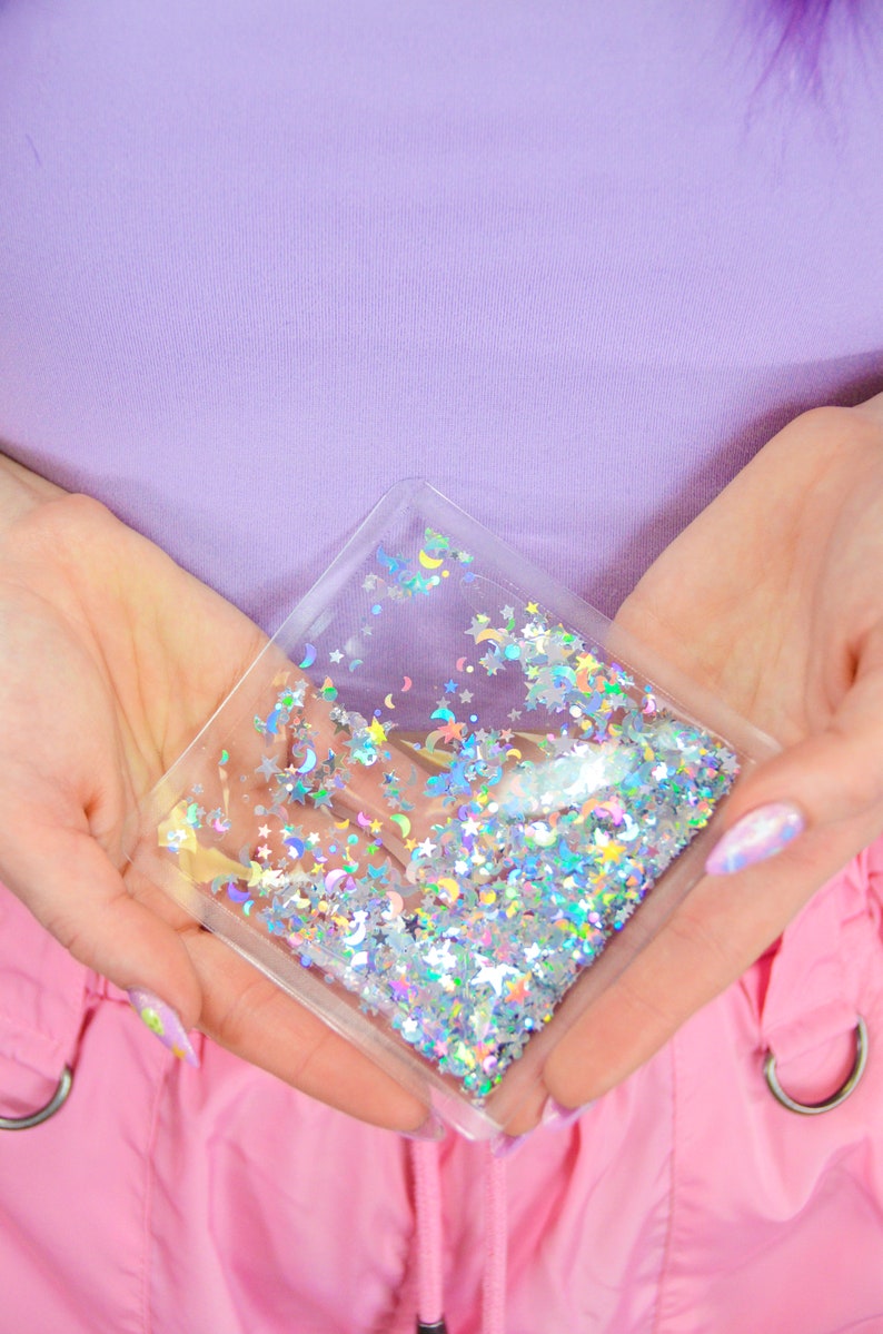 May include: A clear plastic pouch filled with iridescent glitter in the shape of stars and crescent moons. The pouch is held in the hands of a person.