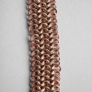 Könnte beinhalten: Ein Armband aus Kupfer- und Goldkettengeflecht mit einem schwarzen Verschluss. Das Armband besteht aus kleinen, ineinandergreifenden Metallringen.