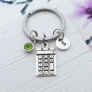 May include: Silver calculator keychain with a green gem, a silver charm with the letter 'S', and a silver key ring.