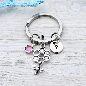 May include: Silver key chain with a pink crystal charm, a honeycomb charm, a bee charm, and a silver charm with the letter "P" on it.