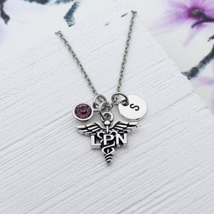 May include: Silver necklace with a medical caduceus charm, a purple crystal charm, and a silver charm with the letter 'S'.