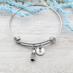 May include: A silver bangle bracelet with a charm of a graduation cap and a charm with the letter K. The bracelet is on a white wooden surface.