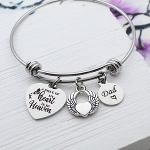 May include: A silver bangle bracelet with three charms. The charms are a heart with the text "A piece of my heart is in Heaven", a heart with angel wings, and a circle with the text "Dad" and a small heart.