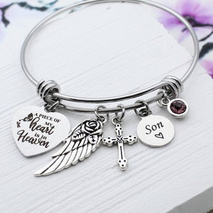 May include: A silver bangle bracelet with charms. The charms include a heart with the text "A piece of my heart is in Heaven", an angel wing, a cross, and a circle with the word "Son" and a heart.