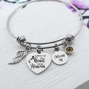 May include: A silver charm bracelet with a heart charm that says "A piece of my heart is in heaven" and a round charm that says "Mom". The bracelet also has a small angel wing charm and a brown crystal charm.