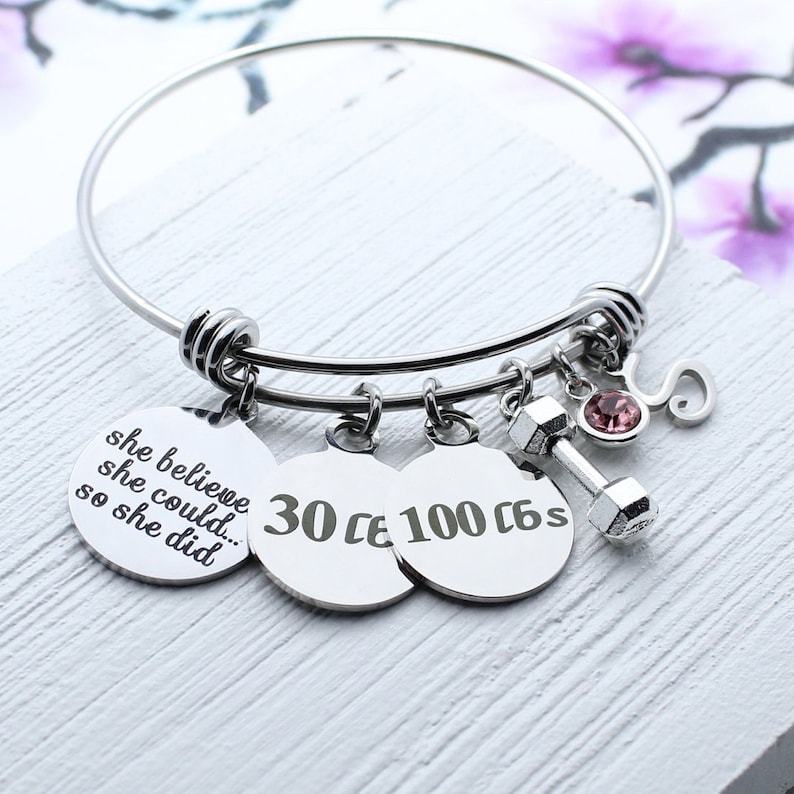 May include: Silver charm bracelet with a dumbbell charm, a pink rhinestone, and three round charms with the text "she believe she could... so she did", "30", and "100 6s".