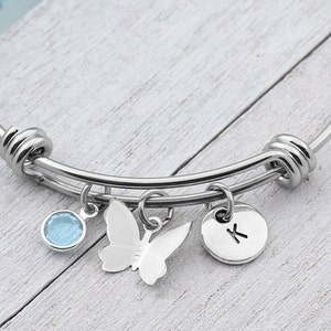 May include: A silver bangle bracelet with a charm of a butterfly, a light blue crystal, and a silver charm with the letter "K" on it.