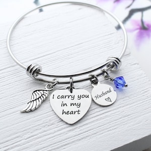 May include: A silver bangle bracelet with charms. The charms include a heart with the text "I carry you in my heart", a round charm with the text "Husband" and a heart, and a small blue crystal charm.