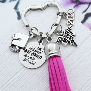 May include: Silver-toned keychain with a heart-shaped ring, featuring charms of a graduation cap, a medical symbol, and a tassel. The keychain includes a charm with the text "She believed she could so she did" and the year "2020". The tassel is a vibrant pink.