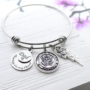 May include: A silver expandable bangle bracelet with three charms. The charms are a round charm with the words "Dad Always in my Heart" engraved on it, a round charm with the United States Army seal, and a silver angel wing charm.