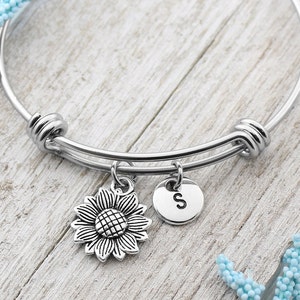 Peut inclure: Un bracelet jonc argenté avec un charme en forme de tournesol et un charme avec la lettre 'S' qui pend.