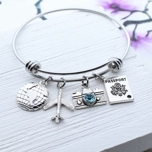 May include: A silver charm bracelet with four charms: a globe, an airplane, a camera with a blue gem, and a passport. The bracelet is on a white wooden surface.