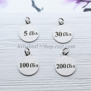 May include: Four stainless steel round charms with attached loops. Each charm has a different weight measurement in pounds: 5 lbs, 30 lbs, 100 lbs, and 200 lbs. The charms are displayed on a light wood-grain surface.