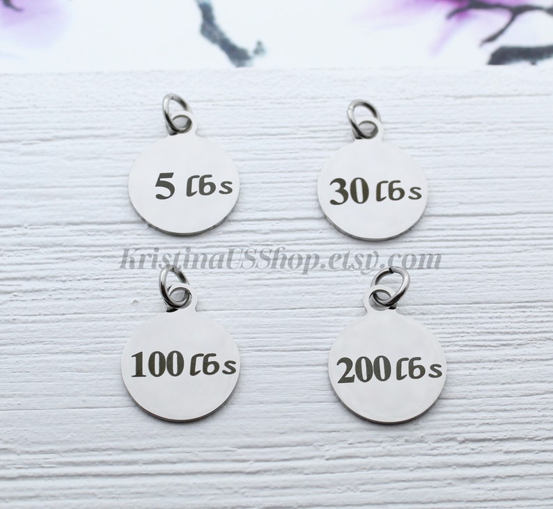 May include: Four stainless steel round charms with attached loops. Each charm has a different weight measurement in pounds: 5 lbs, 30 lbs, 100 lbs, and 200 lbs. The charms are displayed on a light wood-grain surface.