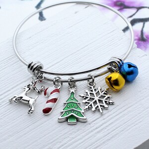 May include: A silver bangle bracelet with five charms: a reindeer, a candy cane, a Christmas tree, a snowflake, and two jingle bells. The bells are gold and blue.