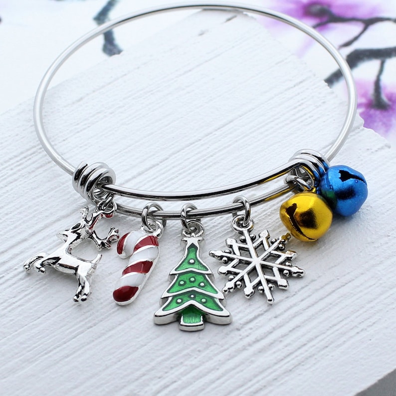 May include: A silver bangle bracelet with five charms: a reindeer, a candy cane, a Christmas tree, a snowflake, and two jingle bells. The bells are gold and blue.