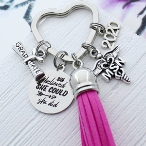 May include: A pink tassel keychain with a silver heart-shaped ring, a silver charm with the text "She believed she could so she did", a silver charm with the text "Graduate", a silver charm with the text "MSN 2020", and a silver charm with a caduceus symbol.
