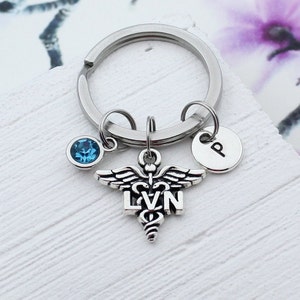 May include: Silver keychain with a medical caduceus charm, the letters "LVN" and a blue crystal charm. The keychain also has a silver charm with the letter "P" on it.