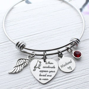 May include: A silver bangle bracelet with charms. The charms include a cardinal, a heart with the text "When cardinals appear your loved one is near", a heart with the text "Husband", and a red crystal.