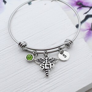 May include: Silver bangle bracelet with a green crystal charm, a silver charm with the medical symbol and the letters "SLP", and a silver charm with the letter "S".