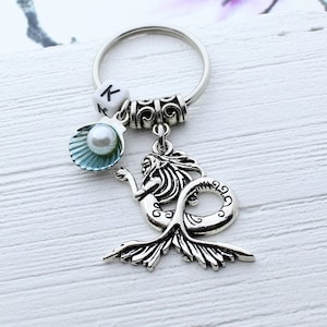 Puede incluir: Llavero de plata con una sirena, un encanto de perla y concha. El llavero también tiene un encanto de plata con la letra K.