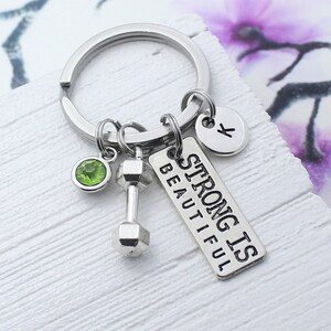 Puede incluir: Llavero de plata con un dije de mancuerna, un dije de cristal verde, un dije de plata con la letra K y una etiqueta de plata que dice "Strong is Beautiful".
