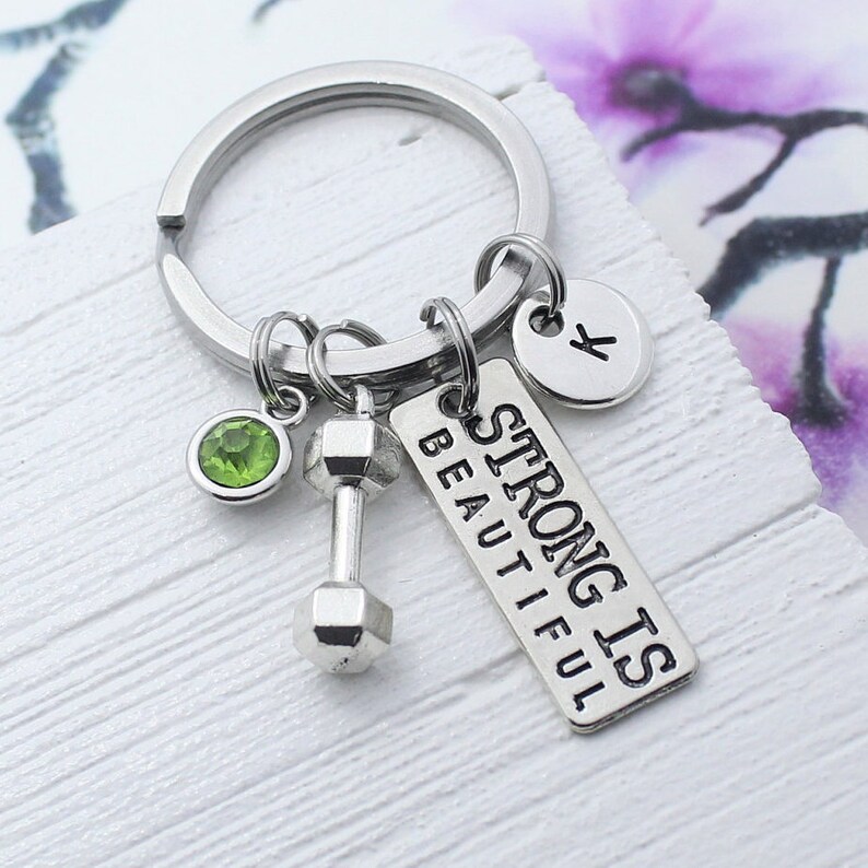 Puede incluir: Llavero de plata con un dije de mancuerna, un dije de cristal verde, un dije de plata con la letra K y una etiqueta de plata que dice "Strong is Beautiful".