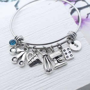May include: A silver charm bracelet with a blue crystal charm and charms shaped like kitchen utensils, including a measuring cup, a whisk, a mixer, and a baking sheet. The bracelet also has a charm with the letter "S" on it.
