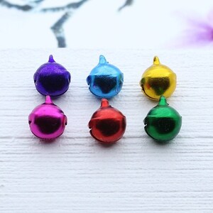 May include: Six small metal bells in different colors: purple, blue, gold, pink, red, and green. The bells are arranged in two rows of three on a white surface.