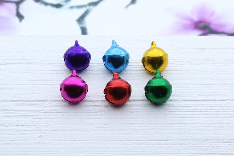 May include: Six small metal bells in different colors: purple, blue, gold, pink, red, and green. The bells are arranged in two rows of three on a white surface.