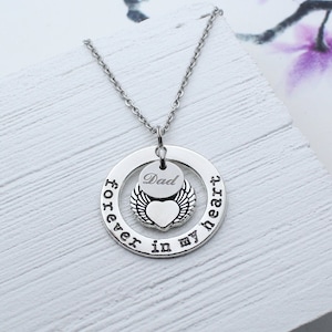 Könnte beinhalten: Silberne Halskette mit einem Herz-Anhänger und einem runden Anhänger, auf dem "forever in my heart" steht. Der Herz-Anhänger hat Flügel und der runde Anhänger hat das Wort "Dad" eingraviert.