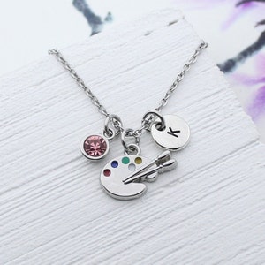 May include: Silver-toned necklace with three charms. One charm is a pink gemstone, another is a small disc with the letter "K", and the third is a painter's palette with colorful paint dots and two paintbrushes.