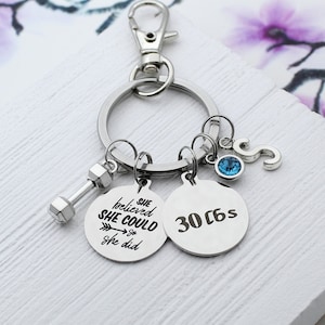 May include: Silver-toned keychain with a dumbbell charm, two round pendants, and a letter "S" charm. One pendant reads "She believed she could so she did." The other pendant says "30 c6s." A blue gem is also attached.