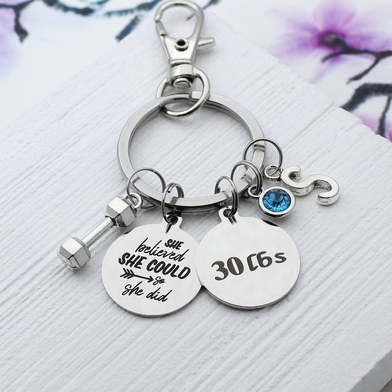 May include: Silver-toned keychain with a dumbbell charm, two round pendants, and a letter "S" charm. One pendant reads "She believed she could so she did." The other pendant says "30 c6s." A blue gem is also attached.