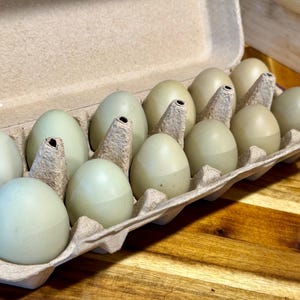 May include: A dozen green eggs in a cardboard egg carton. The eggs vary in shades of green and are nestled in individual compartments. The carton is open, revealing the eggs against a wooden surface.