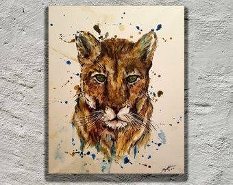 May include: Watercolor painting of a cougar's face with green eyes. The background is a light beige with blue and brown watercolor splatters.